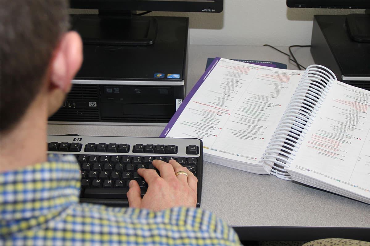 An individual sits at a computer, typing on the keyboard, with a book of medical coding information open on the desk next to them